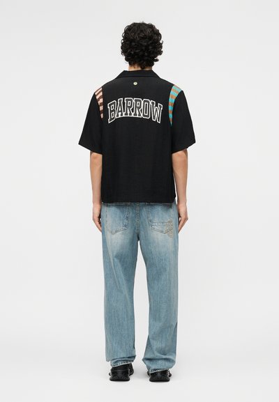 Άτομο με σγουρά μαλλιά στέκεται προς τα πίσω φορώντας μαύρο κοντομάνικο πουκάμισο με το κείμενο "BARROW" και ανοιχτό μπλε τζιν με μαύρα παπούτσια.