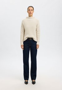 Beige gerippter Pullover mit hohem Kragen, kombiniert mit dunkelblauen geraden Jeans und schwarzen flachen Schuhen. Einfaches, minimalistisches Design.