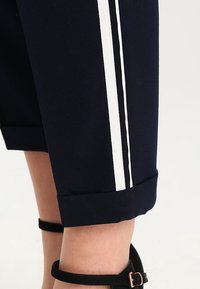 Granatowe spodnie 3/4 z białymi paskami po bokach, z mankietami i gładką fakturą materiału, w zestawieniu z czarnymi butami na kostkę z paskiem.