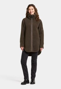 Braune Fleecejacke mit hohem Kragen, durchgehendem Reißverschluss und langen Ärmeln. Über dunklen Hosen und schwarzen Knöchelstiefeln getragen.