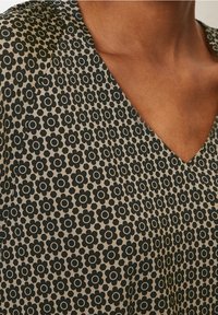 Top marrón con cuello en V que presenta un patrón floral negro repetido. El tejido parece suave con un ligero brillo. Detalle del escote curvado visible.