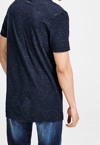 Námořnická modrá tričko s krátkým rukávem a tečkovaným vzorem. Kulatý výstřih, volný střih a měkká textura. Nosené s modrými džíny.