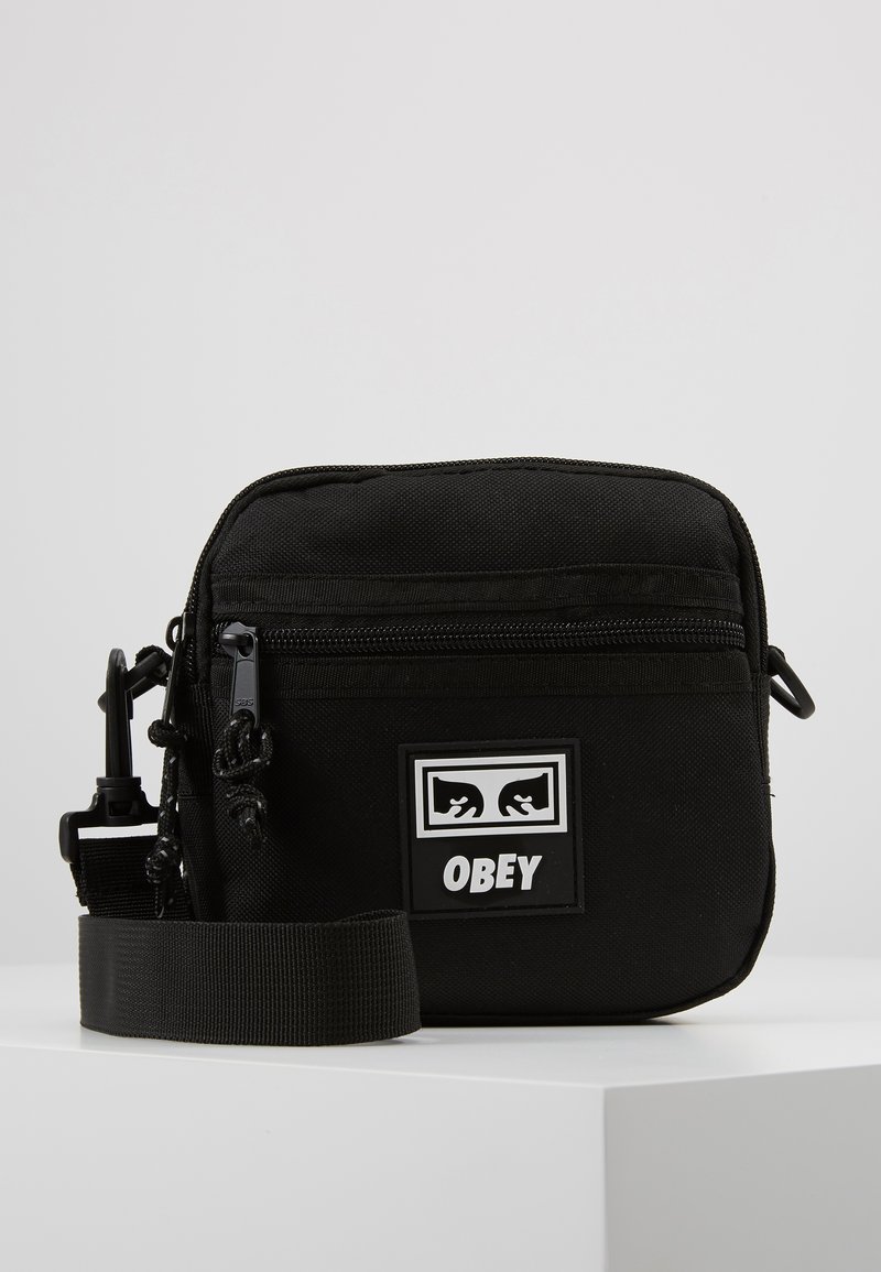 Czarna torba na ramię wykonana z trwałego materiału, z zewnętrzną kieszenią na zamek, regulowanym paskiem oraz prostokątną naszywką z logo "OBEY."