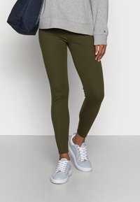 Olive green legginsy z rozciągliwego materiału, w połączeniu z jasnoniebieskimi trampkami z białymi podeszwami i sznurówkami oraz szarym swetrem.