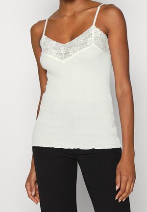 Kobieta ubrana w biały top na ramiączkach z koronkową lamówką w połączeniu z czarnymi spodniami, stojąca na tle gładkiego, jasnego tła.