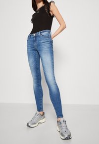 Jeans ajustados de denim azul medio, con un diseño clásico de cinco bolsillos. Llevados con una blusa negra con detalles de encaje y zapatillas grises.