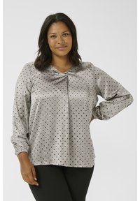 Blouse grise à pois en tissu doux et satiné, avec un col en V, des manches froncées et un détail de pli central. Texture lisse et coupe décontractée.