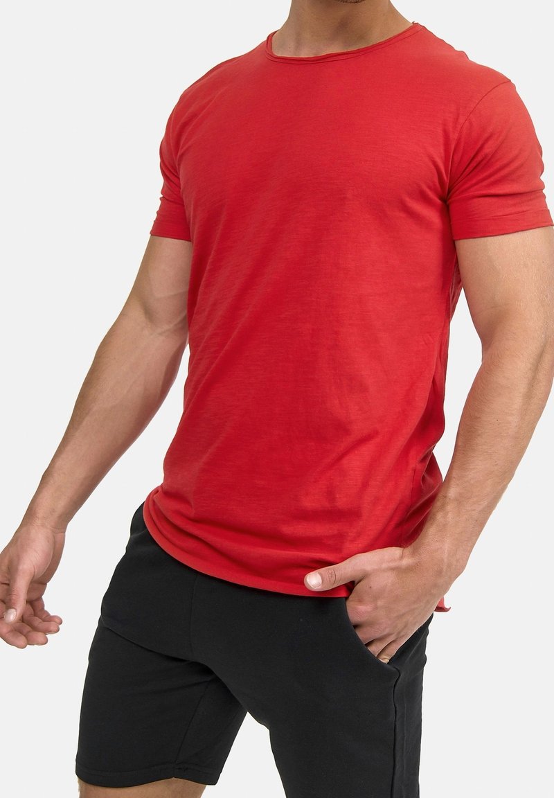 Homme portant un t-shirt rouge uni à manches courtes avec un short noir, une main dans la poche, sur un fond clair uni.