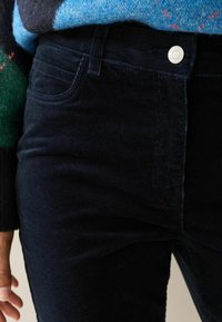 Tmavě modré kordové kalhoty s hladkou texturou, designem s pěti kapsami a kovovým knoflíkem na pase, částečně zakryté svetr.