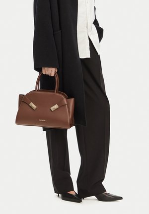 Persona vistiendo pantalones negros de pierna ancha, abrigo negro, camisa blanca y zapatos negros puntiagudos, sosteniendo un bolso de mano de cuero marrón por las asas.