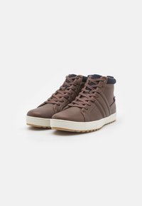 Zapatos altos marrones de cuero liso con suela de goma blanca. Presentan un cuello de tela negra y cordones en contraste con un acabado texturizado.