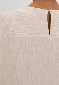 Ljus beige texturerad blus med vertikala ränder, med en nyckelhålsknäppning i ryggen och rund halsringning. Mjuk, andningsbar material.