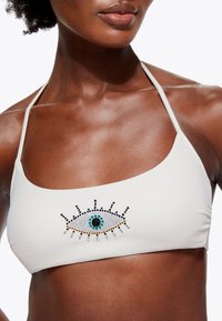 Top bikini bianco con scollatura quadrata, caratterizzato da un disegno di un occhio realizzato con strass neri, turchesi e trasparenti sul davanti. Spalline sottili.