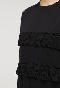 Top in tessuto nero caratterizzato da tre strati orizzontali di ruffles plissè, separati da inserti in pizzo, con scollatura rotonda e maniche corte.