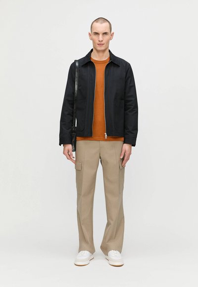 Veste zippée noire par-dessus une chemise orange, pantalon cargo beige et sneakers blanches. La veste a un col et deux poches à l'avant.