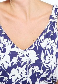 Femme portant un haut sans manches à motifs floraux bleu et blanc avec une bretelle nouée sur l'épaule droite.