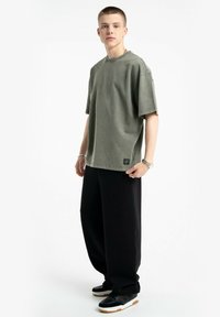 Zielona, oversize bawełniana koszulka z okrągłym dekoltem, podwiniętymi rękawami i małą naszywką z logo; w połączeniu z czarnymi spodniami o szerokich nogawkach i trampkami.