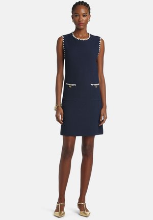 Tamsiai mėlyna rankovių neturinti suknelė iš tekstūruotos medžiagos, su baltais apvadais aplink iškirptę ir kišenes, puošta auksiniais mygtukais.