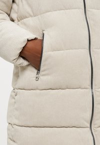 Beige kordfløjljakke med et quiltet design, der har en lynlåslomme og en sort lynlås foran. Blød tekstur og varm udseende.