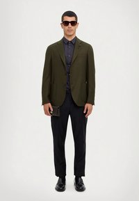 Oliwkowozielona, dopasowana marynarka na ciemnej koszuli, czarne spodnie i buty. Model trzyma mały czarny portfel. Kwadratowe okulary przeciwsłoneczne dopełniają look.