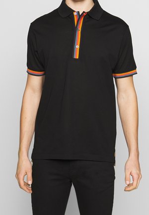 Mann trägt ein schwarzes Poloshirt mit regenbogenfarbenem Streifen am Knopfleistenrand und an den kurzen Ärmeln, kombiniert mit schwarzen Hosen.