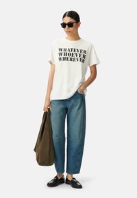 T-shirt en coton blanc avec un texte noir en gras "PEU IMPORTE QUI, PEU IMPORTE OÙ", associé à un pantalon en denim bleu, des chaussures noires et un sac marron.