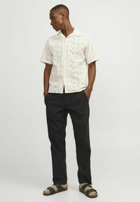 Camisa blanca de manga corta con un diseño estampado, combinada con pantalones oscuros. La camisa tiene cuello y un corte relajado. Sandalias marrones completan el look.