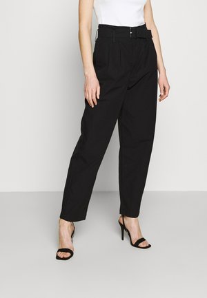 Mujer con pantalones negros de cintura alta con cinturón, combinados con sandalias negras de tacón alto y una camiseta blanca sin mangas.