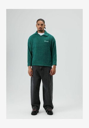 Turkooise fleece pullover met een kraag, lange mouwen en een wit logo op de borst. Gecombineerd met zwarte wijde jeans en zwarte schoenen.