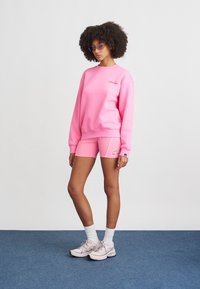 Žiarivá ružová mikina s rebrovanými manžetami, zodpovedajúce ružové šortky s bielymi akcentmi a svetlé tenisky na modrom koberci.