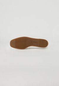 Sneaker blanc avec une tige texturée de couleur crème et une semelle en caoutchouc marron présentant des stries horizontales pour l'adhérence. Marque visible sur la semelle.