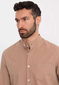 Chemise boutonnée beige clair avec un col mandarin, une poche poitrine unique, et un tissu texturé. Dotée de boutons blancs et d'une coupe décontractée.