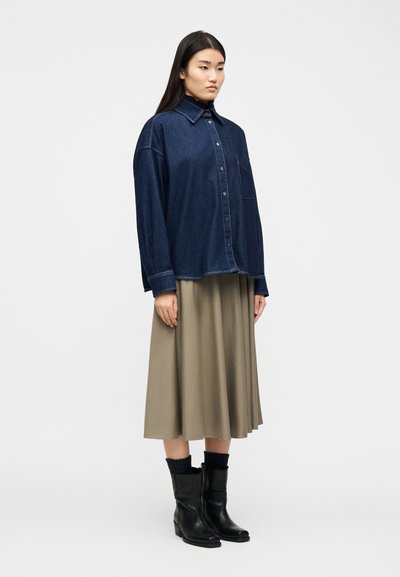 Γυναίκα που φοράει σκούρο τζιν πουκάμισο oversized, μπεζ μάδι φούστα, μαύρες κάλτσες και μαύρα μποτάκια αστραγάλου, στέκεται μπροστά σε απλό λευκό φόντο.