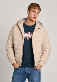 Chaqueta de plumas beige con capucha, presenta un textura acolchada y puños acanalados. Lleva encima una camiseta de color marino con un gráfico de la bandera británica. Jeans visibles.