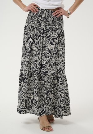 Femme portant une jupe longue à motif paisley noir et blanc avec une ceinture froncée et des sandales beiges à bouts ouverts, mains posées sur les hanches.