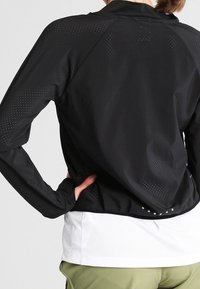 Černá sportovní bunda s dlouhými rukávy a špičatým límcem, s perforovanými detaily na zadní straně a volným střihem přes bílou košili.