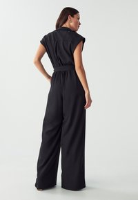 Zwarte jumpsuit van soepele stof met korte mouwen, een ceintuur in de taille en brede pijpen. Bevat een kraag en naden op de schouders.