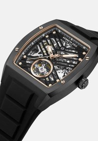 Schwarze rechteckige Uhr mit einem Skelett-Zifferblatt, goldenen Akzenten und einem strukturierten Gummiband. Sie zeigt sichtbare Zahnräder und eine charakteristische Krone.