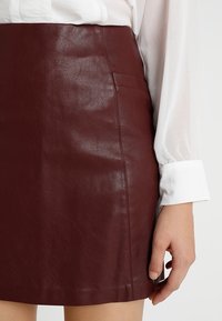 Femme portant une jupe en cuir marron foncé et un chemisier blanc à manches longues, la main reposant à côté de la jupe.