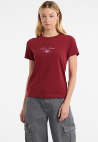 Červené bavlnené tričko s okrúhlym výstrihom a krátkymi rukávmi; na prednej strane je vyšívané logo "Guess Jeans" v kontrastnej farbe.