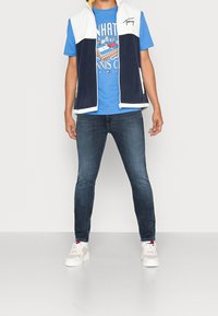 Marinblå fleeceväst med vita detaljer, dragkedja fram, kombinerad med en blå grafisk t-shirt, mörka jeans och ljusa sneakers.