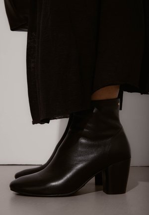 Bottes en cuir noir à talons blocs portées avec une longue jupe noire sur un fond clair.