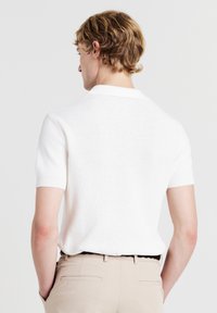 Polo a maniche corte, bianco, tessuto testurizzato, colletto a coste, vestibilità slim. Indossato con pantaloni beige, visione della parte posteriore e delle spalle.