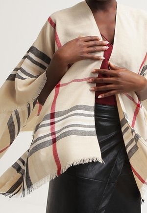 Osoba o ciemnej karnacji ubrana w beżowy szal w czerwone i szare paski, na czerwonym topie i czarnej skórzanej spódnicy, trzymająca szal zaciśnięty.