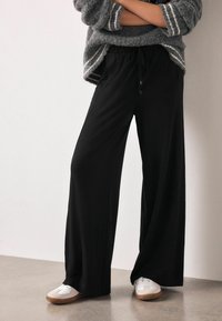 Schwarze weit geschnittene Hose mit elastischem Bund und Kordelzug. Kombiniert mit weißen Turnschuhen und einem grauen Strickpullover mit Streifen.