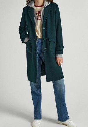 Personne portant un manteau boutonné vert foncé par-dessus un sweat à capuche gris, un t-shirt beige imprimé, un jean bleu et des baskets blanches, debout les mains détendues.