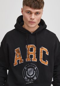 Sudadera negra con capucha que presenta las letras "ARC" en un texto naranja desgastado, con elementos gráficos en blanco y cordones.