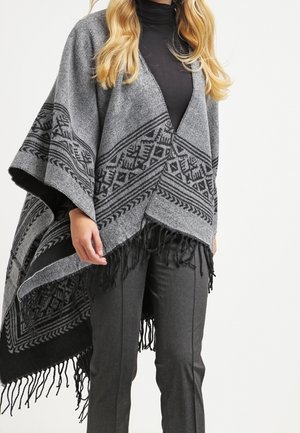 Femme portant un poncho gris à motifs avec des franges par-dessus un col roulé noir et un pantalon gris foncé sur un fond uni.