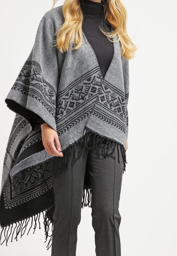 Femme portant un poncho gris à motifs avec des franges par-dessus un col roulé noir et un pantalon gris foncé sur un fond uni.