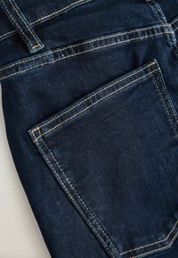 Tmavomodré rifle z denimu s výrazným žltým prešívaním a viditeľným zadným vreckom na svetlom pozadí.